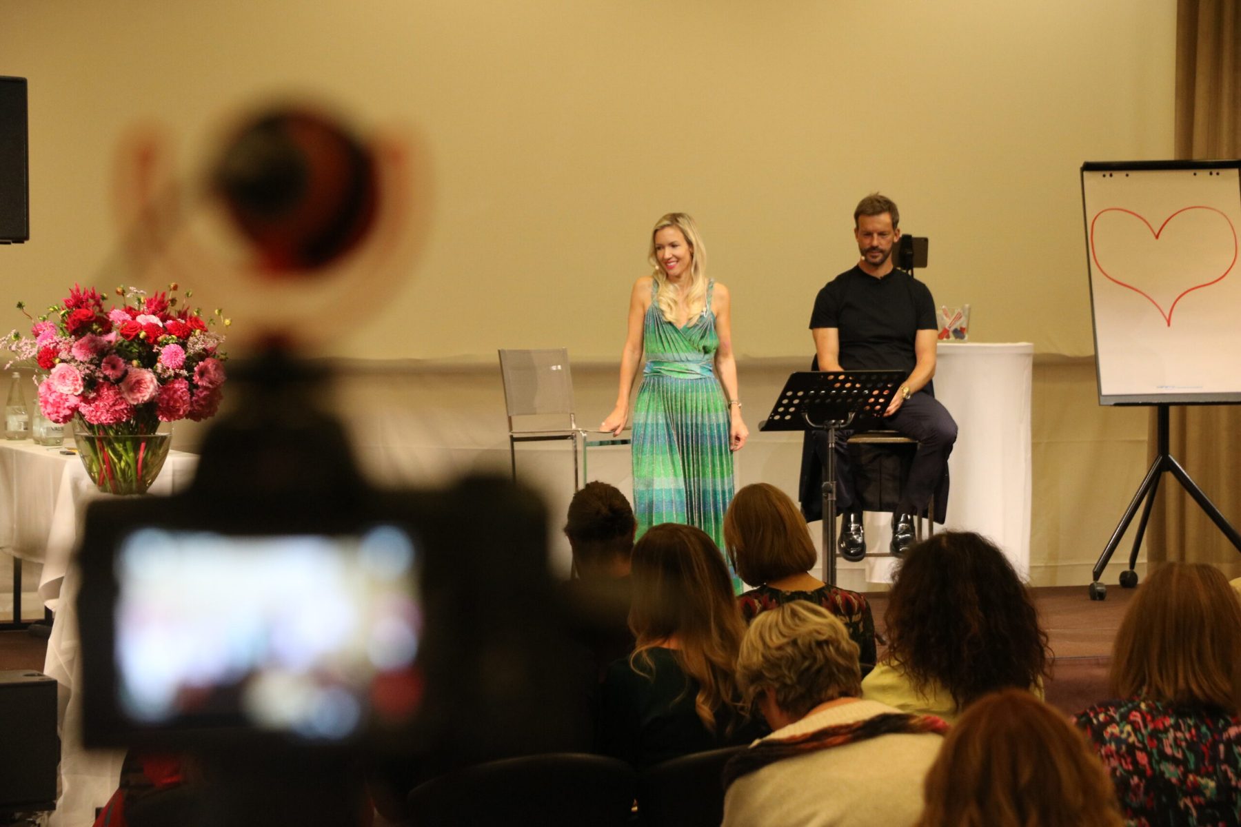 A live ceremony being filmed for streaming, the couple on stage in front of a seated audience and floral arrangements control on a laptop with a stream deck